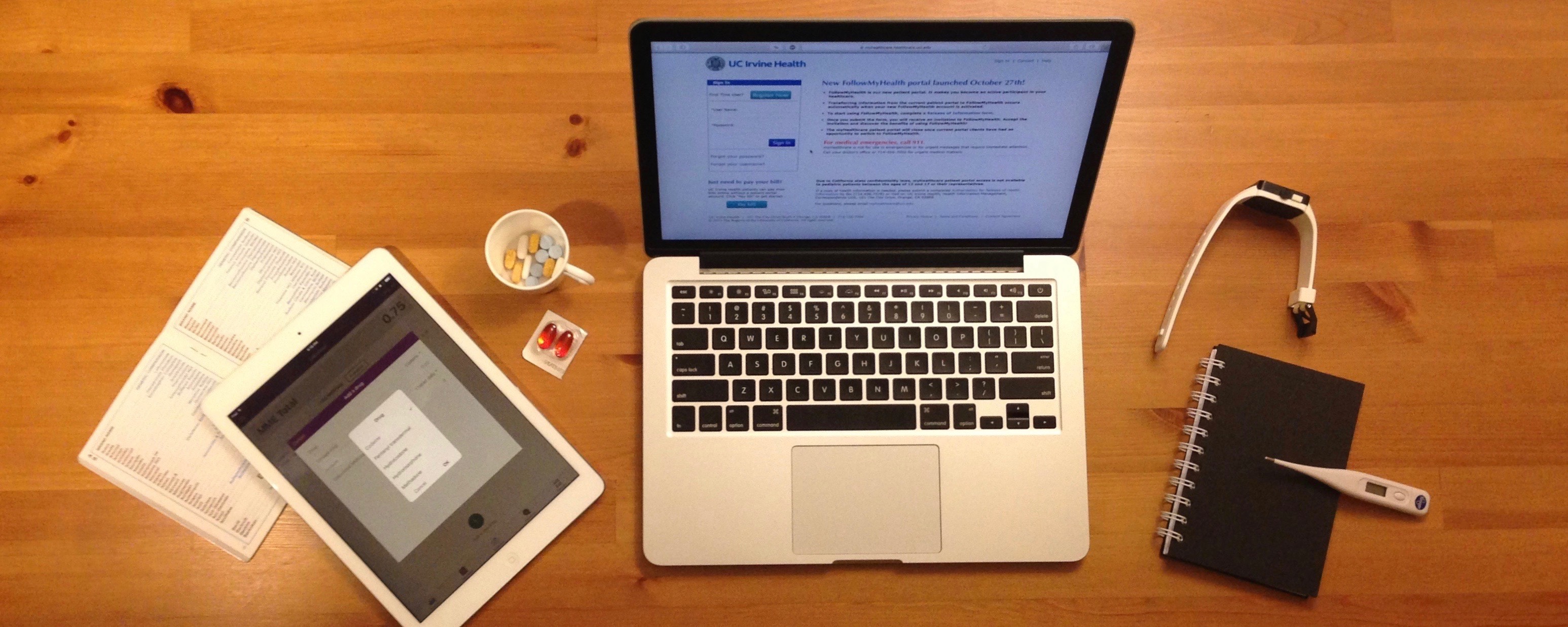 Left to right: A brand-to-generic handbook, a dose calculator on an iPad, assorted medicines, a laptop displaying a patient portal login screen, a fitness band, a patient diary, and a thermometer.