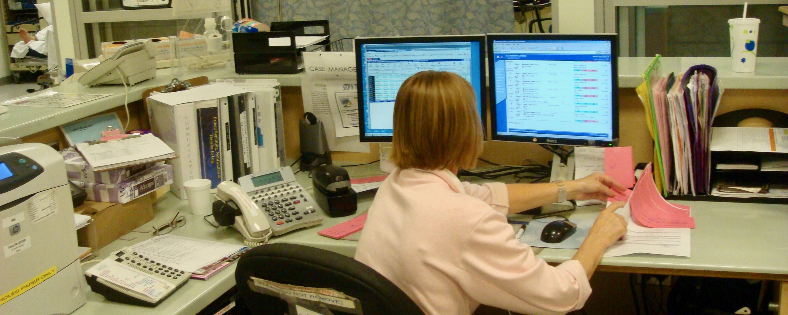 A case worker sits at an emergency department desk cluttered with many artifacts, including paper, desktop displays, and a telephone.